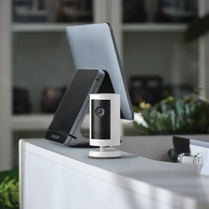 Ring Außenkamera Plus (Plug) - 2.048 x 1.536 Pixel (2K-HD), Weiß - Eine Ring Sicherheitskamera, die auf einer weißen Oberfläche neben einem Laptop und einem Smartphone steht, bietet Sicherheit für Ihr Zuhause.
