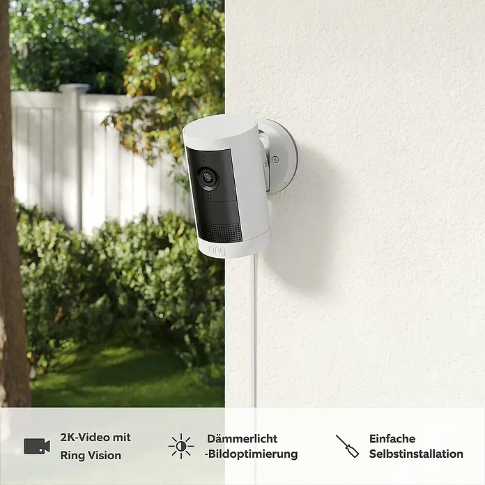 Ring Außenkamera Plus (Plug) - 2.048 x 1.536 Pixel (2K-HD), Weiß - Eine Ring Spotlight Cam ist an einer weißen Wand montiert, mit einem Garten im Hintergrund.