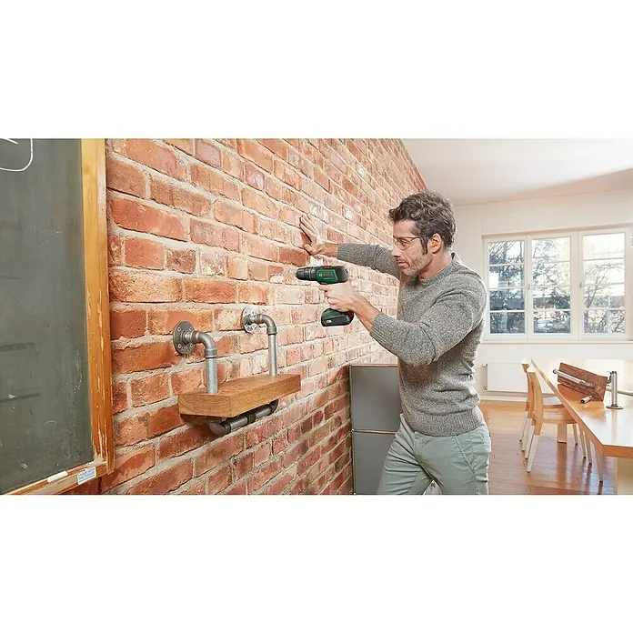 Bosch Power for All 18V Akkuschrauber UniversalImpact 18V - 18 V, 2 Akkus, 1,5 Ah, 241 -tlg. - Ein Mann bohrt mit einer grünen Bosch-Bohrmaschine in eine Ziegelwand, um ein Regal mit Metallrohrstützen zu befestigen.