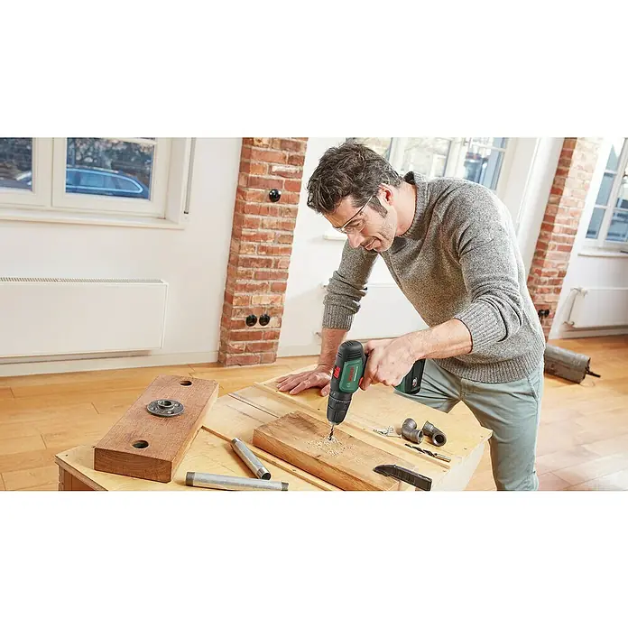 Bosch Power for All 18V Akkuschrauber UniversalImpact 18V - 18 V, 2 Akkus, 1,5 Ah, 241 -tlg. - Mann bohrt mit Akkubohrer Löcher in ein Holzbrett. Die Bohrmaschine ist schwarz und grün. Die Person trägt eine Schutzbrille.