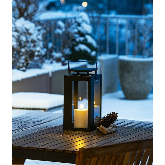 LED-Laterne Lucido - 15 x 15 x 31 cm, Schwarz - Eine stimmungsvolle Laterne mit Kerze und Zapfen auf einem Holztisch im winterlichen Garten, die eine warme Atmosphäre erzeugt.