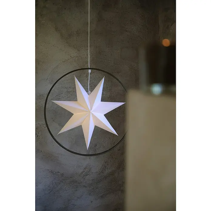 Markslöjd Pendelleuchte Speranza - Ø x H: 50 x 17 cm, Weiß/Grau - Eine leuchtende Papierstern-Lampe hängt an einem weißen Kabel vor einer grauen Wand und einem unscharfen Objekt.