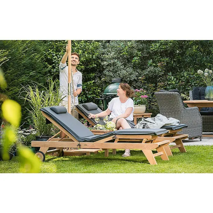 Kai Wiechmann Liegenauflage - Sonnenliege, Hochlehner, Relaxchair, L x B x H: 49 x 36 x 6 cm, Polyacryl, Anthrazit - Ein Mann stellt einen Sonnenschirm auf, während eine Frau auf einer Outdoor-Sonnenliege entspannt. Bequeme Gartenmöbel in Szene gesetzt.