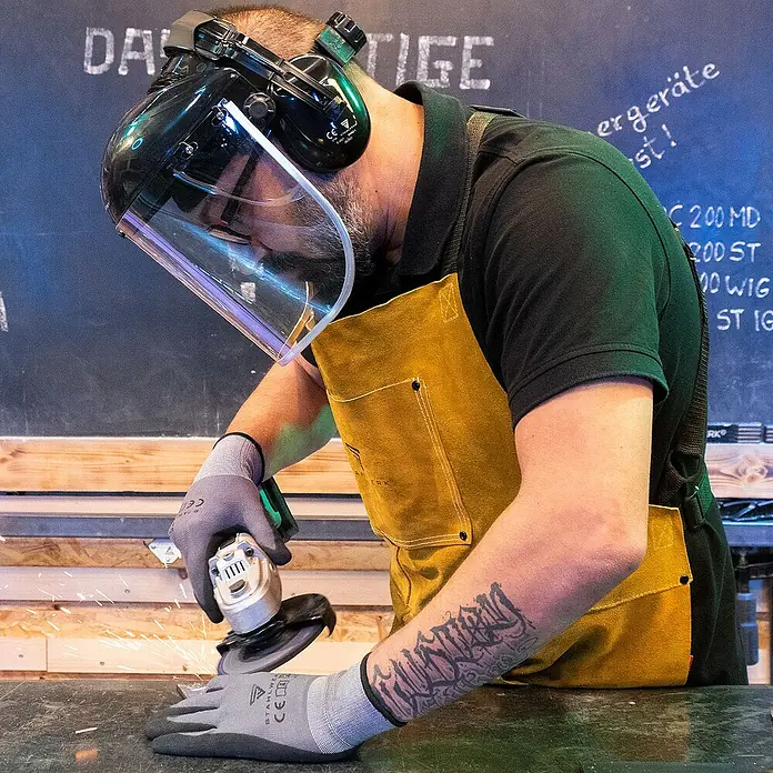 STAHLWERK Schutzhelm 2 x Gesichtsschutz mit Visier -  - Ein Mann mit Schutzhelm und Schürze schleift Metall mit einem Winkelschleifer.