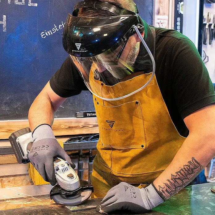 STAHLWERK Schutzhelm Gesichtsschutzschild 3er Set -  - Nahaufnahme eines Mannes, der mit einer STAHLWERK-Schutzmaske und -Schürze Metall schleift.