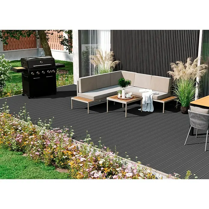 Kovalex WPC-Terrassendiele Exklusiv nach Maß - 600 x 14,5 x 2,6 cm, Steingrau - Elegantes Terrassenensemble mit Grill und Lounge-Möbeln auf dunklem Holzboden, umgeben von blühenden Blumen, die eine einladende Außenatmosphäre schaffen.