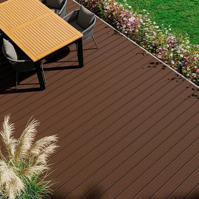 Kovalex WPC-Terrassendiele Exklusiv nach Maß - 300 x 14,5 x 2,6 cm, Braun - Detailaufnahme einer Holzterrasse mit Esstisch und Stühlen, umgeben von Blumen und Rasen. Die Terrasse ist braun und hat eine feine Holzmaserung.