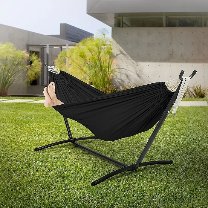 ML-Design Hängematte XXL Hängesessel Schwarz mit Tragetasche und Ständer für Garten/Balkon - L x B: 255 x 99 cm, Stahl, Schwarz, Passend für: Erholung, Lesen, Entspannen - Bild einer schwarzen Hängematte mit Gestell auf einem Rasen vor einem modernen Haus. Die Hängematte lädt zum Entspannen ein.