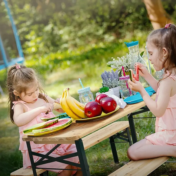 ML-Design Kinder-Picknicktisch Garten Sitzgruppe für Kinder, Braun, Tannenholz, massiv, mit 4 Plätzen - Braun, Dunkelgrün, Holz - Zwei Mädchen sitzen an einem Picknicktisch mit Wassermelone, Bananen und Äpfeln. Getränke und Blumen schmücken den Tisch für ein entspanntes Picknick.