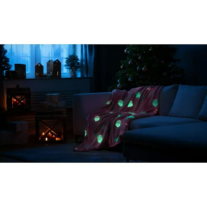 Decke Cherry Xmas - Glow in the Dark - Rot, 180 x 140 cm, 100 % Polyester - Ein rosa Überwurf mit leuchtenden Weihnachtsmannmotiven liegt auf einem Sofa neben einem geschmückten Weihnachtsbaum.