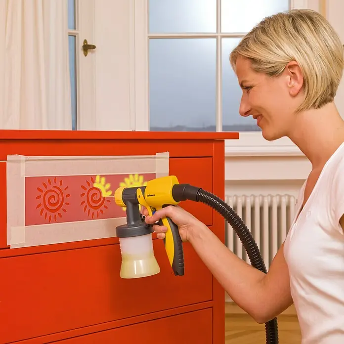 Wagner Sprühaufsatz Wood & Metal EXTRA Detail - Passend für: Wagner Farbsprühsystem Universal Sprayer & Wall Sprayer, Geeignet für: Dünnflüssige lösemittel- & wasserbasierte Materialien - Eine Frau verziert eine Kommode mit einer Farbspritzpistole von Wagner, die gelb und schwarz ist, um sonnenförmige Muster zu malen.