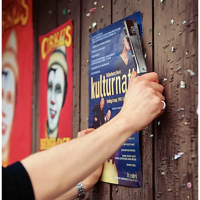 Rapid Hammertacker R19 - Klammertiefe: 4 mm - 6 mm, Geeignet für: Feindrahtklammer Typ 53 - A person staples a "Kulturnatt" poster to a wooden wall with circus posters already attached.