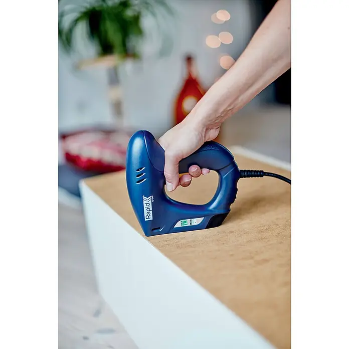 Rapid Kombi-Elektrotacker E-TAC - Klammertiefe: 6 mm - 14 mm, Länge Nagel: 15 mm - Nahaufnahme eines Rapid E-Tac Elektrotackers in Aktion, gehalten von einer Hand auf einer Holzoberfläche, im Hintergrund verschwommene Dekoration.