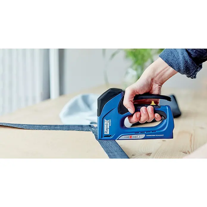 Rapid Handtacker ALU953 - Klammertiefe: 6 mm - 14 mm, Geeignet für: Klammern Typ 53 - Eine Hand benutzt einen blauen Rapid ALU953 Tacker, um ein Textilband auf einem Holztisch zu befestigen. Ideal für Polsterarbeiten und DIY-Projekte.