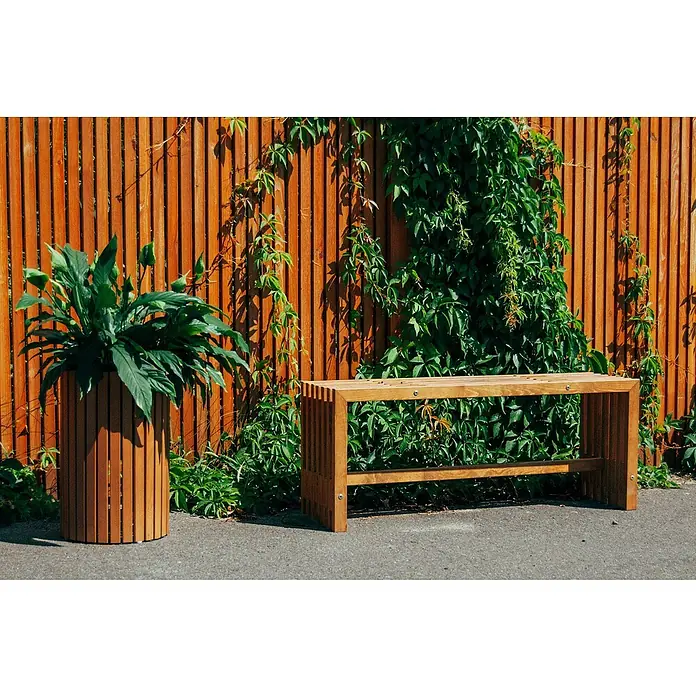 Andrewex Pflanzsäule rund Quadra - Außenmaß (Ø x H): 34 x 40 cm, Holz, Kiefer - Elegante Holzbank und passender Pflanzkübel aus Holzlatten vor einer Holzwand mit Grünpflanzen, die eine ruhige und natürliche Atmosphäre schaffen.