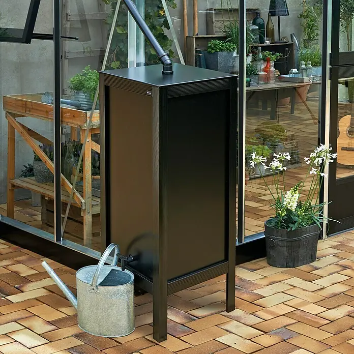 Juliana Regenspeicher - 46 x 46 x 127,7 cm, Schwarz - Eine schwarze Regentonnenanlage mit einer Gießkanne davor, die in einem Garten mit Pflastersteinen und Gewächshaus steht.