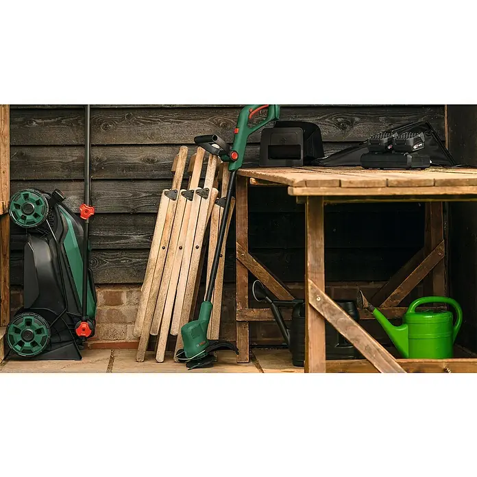 Bosch Power for All 18V Akku-Rasenmäher ROTAK18V2-38 - Ohne Akku, Schnittbreite: 38 cm - Eine Rasenmäher, ein Rasentrimmer, ein Tisch und Regale sind zu sehen. Auf dem Tisch liegen Akkus und eine Tasche.
