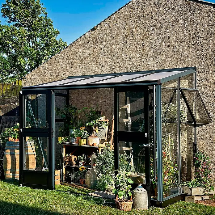 Juliana Gewächshaus Veranda - Außenmaß inkl. Dachüberstand (B x T): 296 x 149 cm, Material Seitenwand: Glas, 3 mm, Anthrazit - Ein Gewächshaus mit geöffneter Tür, gefüllt mit Pflanzen und Gerätschaften, vor einer Hauswand.