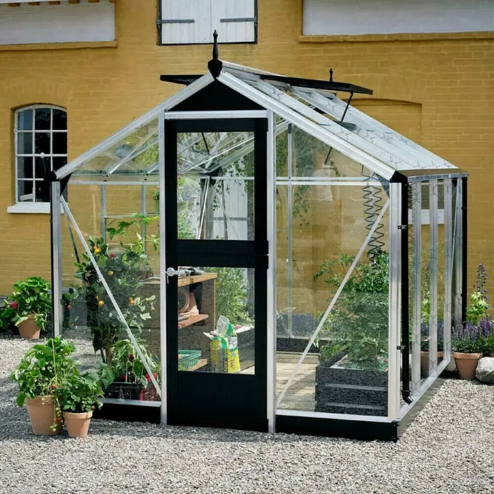Juliana Gewächshaus Compact - Außenmaß inkl. Dachüberstand (B x T): 224 x 295 cm, Material Seitenwand: Glas, 3 mm, Silber/Schwarz - Ein Gewächshaus mit Pflanzen darin steht vor einem gelben Backsteingebäude, umgeben von Kies.