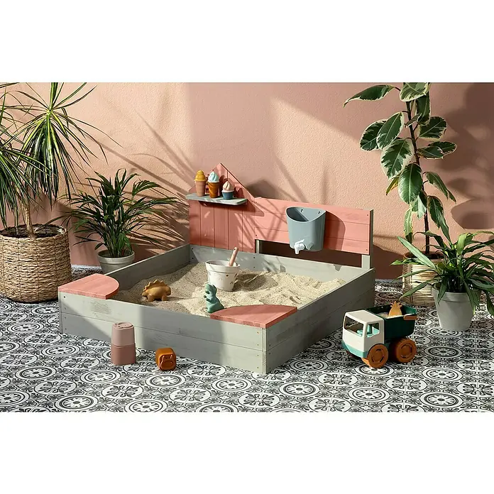 Muddy Buddy Sandkasten Cozy House - 100 x 100 x 60 cm, Holz, Warmgrau/Puderrosa - Helle Aufnahme eines modernen Sandkastens mit Spielzeug, Pflanzen und dekorativem Bodenmuster für ein kindgerechtes Ambiente.