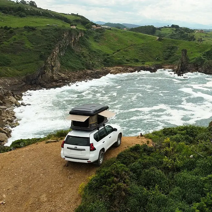 Tagle Outdoor Tienda de campaña para automóvil Hossegor + - Altura: 75 cm, Específico para: 2 personas, Apto para: 2 personas, Escalera telescópica - Un SUV blanco con una tienda de techo negra está aparcado en una costa con un paisaje escénico de fondo.