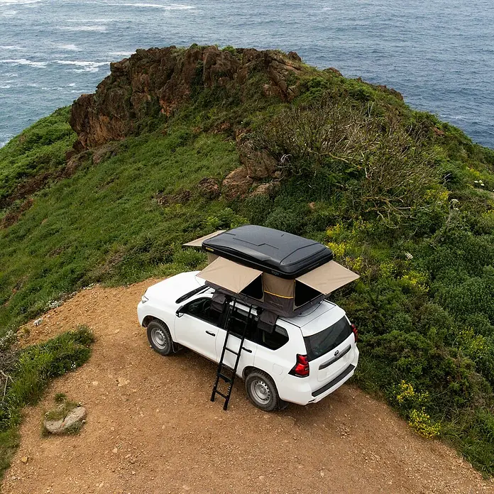 Tagle Outdoor Tienda de campaña para automóvil Hossegor + - Altura: 75 cm, Específico para: 2 personas, Apto para: 2 personas, Escalera telescópica - Un SUV blanco con una tienda de techo Thule instalada en un acantilado con vistas al mar.