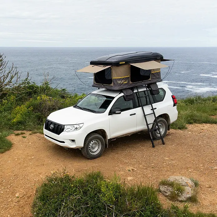 Tagle Outdoor Tienda de campaña para automóvil Hossegor + - Altura: 75 cm, Específico para: 2 personas, Apto para: 2 personas, Escalera telescópica - Un SUV blanco con una tienda de techo "Togile", situado en la cima de una colina con vistas al mar.