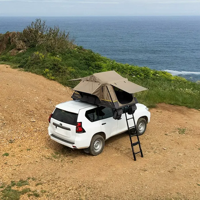 Tagle Outdoor Tienda de campaña para automóvil Somo Lite - Altura: 100 cm, Específico para: 2 personas, Apto para: 2 personas, Techo corredizo - Imagen de un coche blanco con una tienda de techo en una colina con vistas al mar. La tienda de techo es marrón y tiene una escalera.