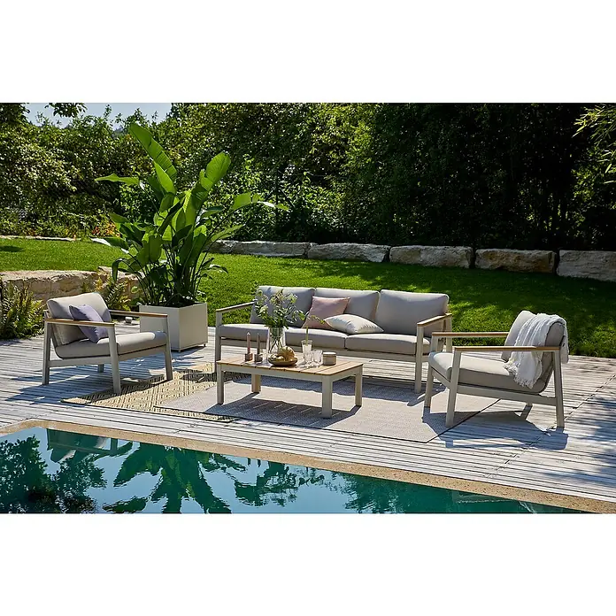 Sunfun Solo Loungemöbel-Set I - 4 -tlg., Aluminium, Olefin, Light-Greige - Graue Gartenmöbel-Garnitur mit Sofa, Sesseln und Tisch neben einem Pool auf einer Holzterrasse, umgeben von Grün.