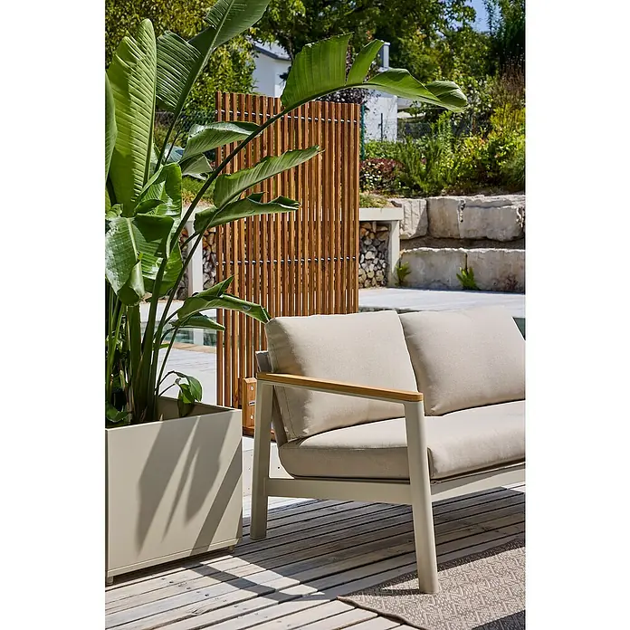Sunfun Raumteiler - 80 x 140 cm, Akazie, Braun - Ein modernes Outdoor-Sofa mit beigen Kissen und einem hellen Holzrahmen steht auf einer Terrasse, umgeben von üppiger Vegetation.