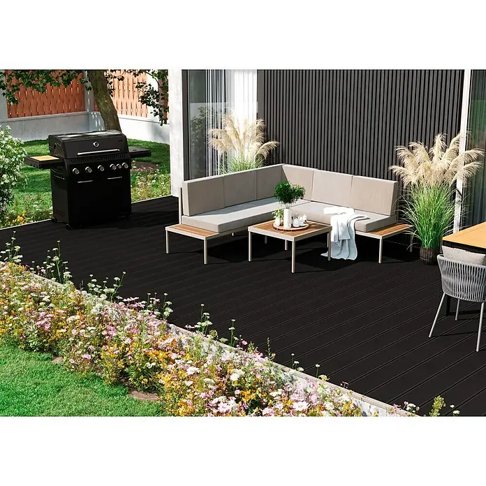 Kovalex WPC-Terrassendiele XL mit Struktur nach Maß - Graubraun, 390 x 19 x 2 cm - Terrassenansicht mit stilvoller Lounge-Garnitur, Grill und blühendem Blumenbeet. Der dunkle Terrassenboden bildet einen eleganten Kontrast zur hellen Umgebung.