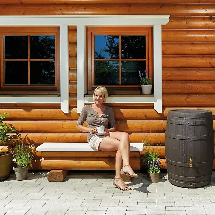 Garantia Classic Line Regenspeicher Barrica - 260 l, Dunkelbraun - Eine Frau sitzt auf einer Bank neben einem dekorativen Regenfass aus Kunststoff in Holzoptik, das Regenwasser stilvoll sammelt.