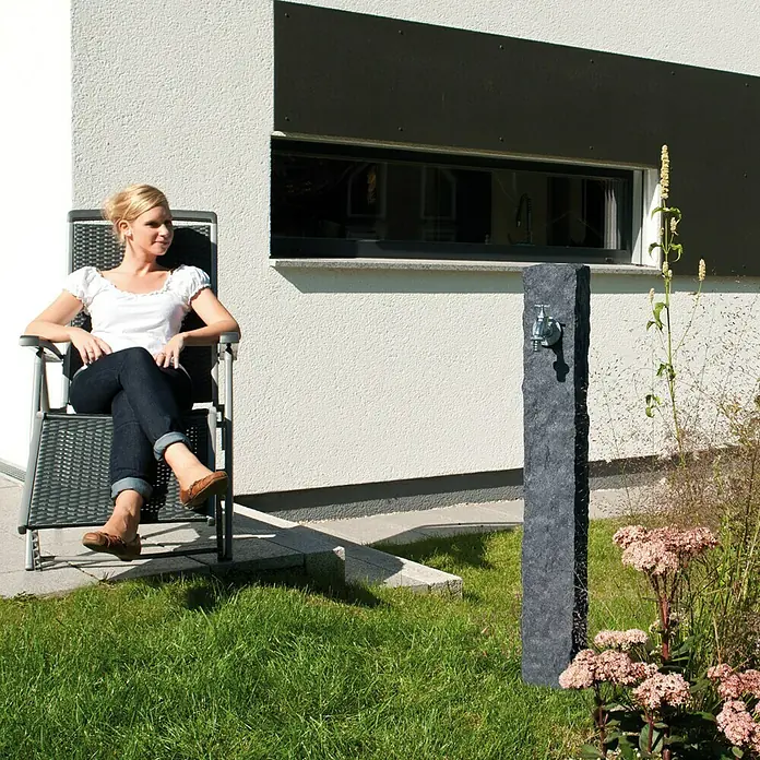 Garantia Wasserzapfstelle Granit - Darkgranite, Höhe: 100 cm - Außenaufnahme einer stilvollen Wasserzapfstelle aus Stein im Garten, neben einer entspannten Frau im Sessel.
