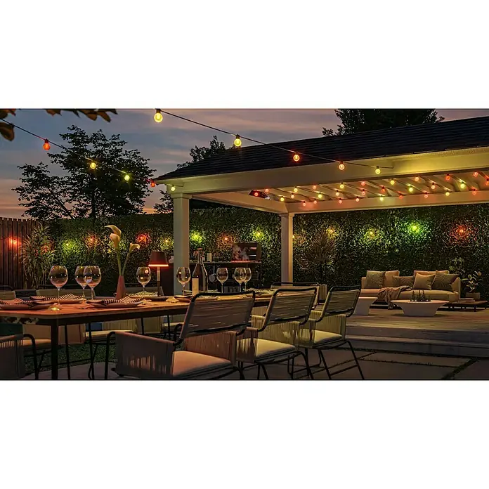 Philips Hue LED-Lichterkette Festavia Globe - 14 m, Anzahl Leuchtmittel: 20 Stk. - Gartenbeleuchtung mit bunten Glühbirnen über einem Tisch und Stühlen, die eine gemütliche Atmosphäre für ein Outdoor-Dinner schaffen.