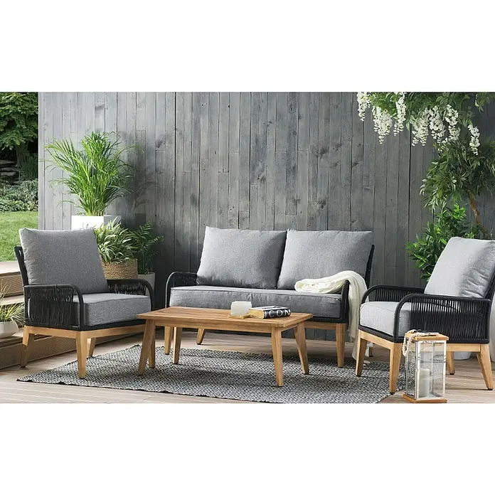 Acamp Loungemöbel-Set Mykonos - 4 -tlg., Akazie, Polyester, Grau - Elegantes Outdoor-Möbelset mit Sofa, Sesseln und Couchtisch auf einem grauen Teppich vor einer grauen Holzwand. Dekorative Pflanzen vervollständigen das Ensemble.