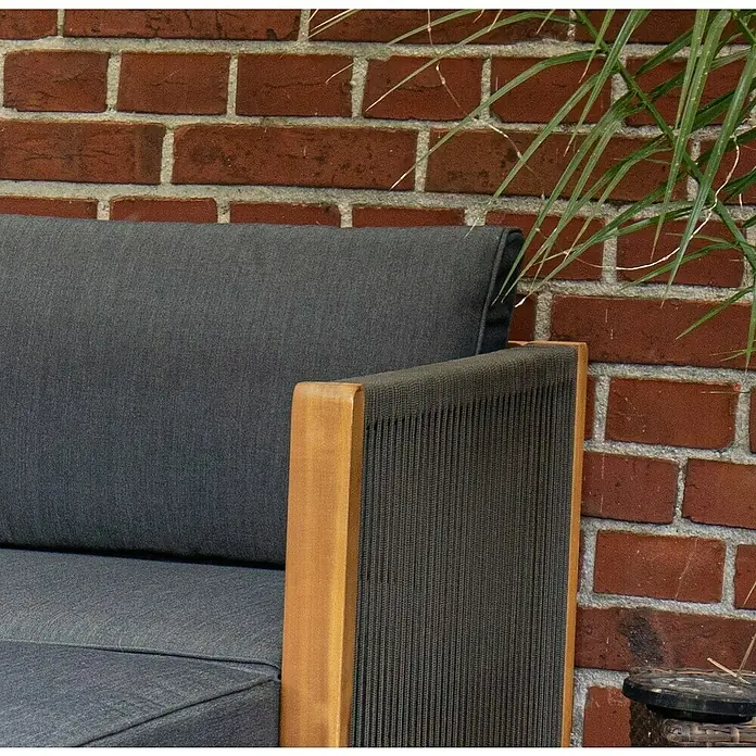 Acamp Loungemöbel-Set Malaga - 3 -tlg., Akazie, Polyester, Braun - Nahaufnahme eines grauen Sofas mit Holzrahmen vor einer Backsteinmauer und einer grünen Pflanze.