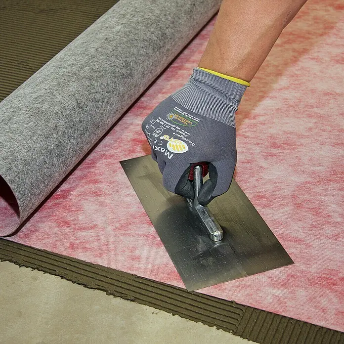 Racofix RF-100 Dichtfolie Meterware - Max. Zuschnittsmaß: 15 m, Breite: 1 m, Als Zuschnitt erhältlich - Ein Handwerker glättet mit einer Kelle eine rote Bauabdichtungsbahn, während eine Rolle graue Abdichtungsbahn bereitliegt.