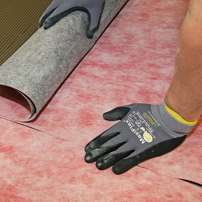 Racofix RF-100 Dichtfolie Meterware - Max. Zuschnittsmaß: 15 m, Breite: 1 m, Als Zuschnitt erhältlich - Ein Arbeiter verlegt eine graue Unterlage auf einer roten Oberfläche mit Maxiflex-Handschuhen für sicheren Halt.