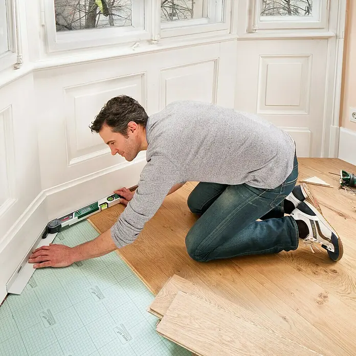 Bosch Goniómetro PAM 220 - Gama de medición: 0 - 220°, Número de niveles de burbuja: 2 ud. - Un hombre colocando un piso de madera con un transportador y un nivel de burbuja en una habitación luminosa.