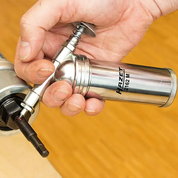 Hazet Fettpresse 2162M - Druck: Max. 69 bar/1.000 psi, Füllmenge: 80 g - Eine Hazet 2162 M Fettpresse wird von einer Hand gehalten, während sie ein Schmiernippel schmiert.