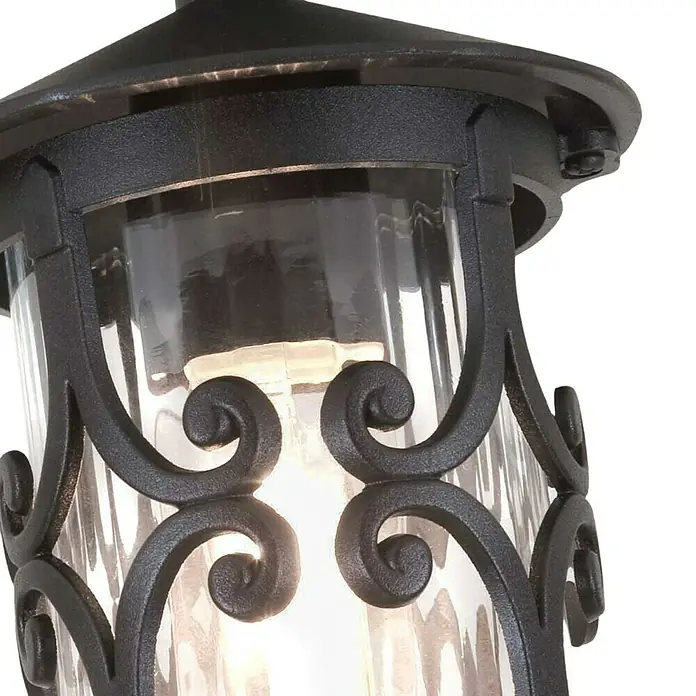 Elstead Außenwandleuchte Hereford - 60 W, 15 x 15 x 26 cm, Schwarz, IP23 - Nahaufnahme einer schwarzen Außenlaterne mit dekorativen Verzierungen und klarem Glas, die ein warmes Licht ausstrahlt.