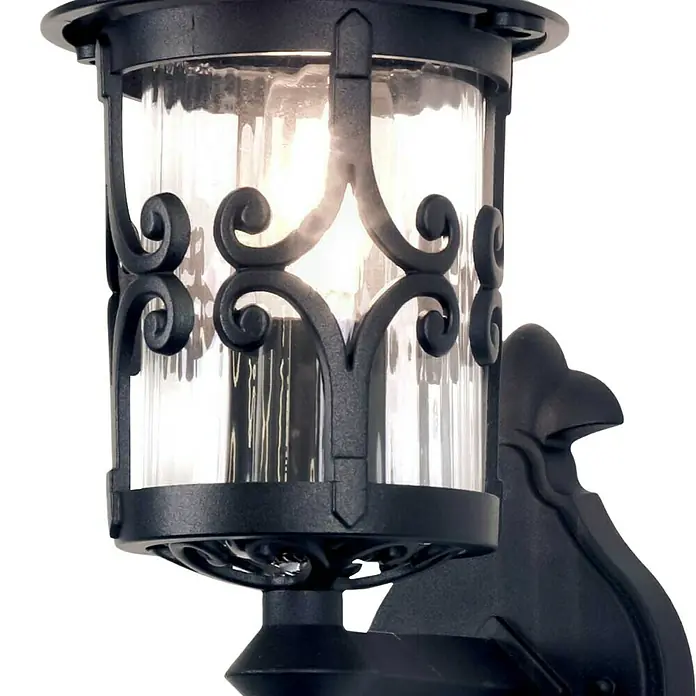 Elstead Außenwandleuchte Hereford - 60 W, 21,5 x 42 x 42 cm, Schwarz, IP23 - Nahaufnahme einer schmiedeeisernen Wandleuchte mit klarem Glaszylinder und dekorativen Scroll-Details. Die Leuchte strahlt warmes Licht aus.