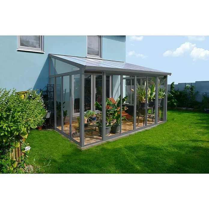 Palram – Canopia Porche acristalado San Remo - An x Pr x Al: 4,25 x 3 x 3,1 m, Gris antracita - Un invernadero con paredes de vidrio y techo transparente está adjunto a una casa y rodeado de exuberante vegetación.