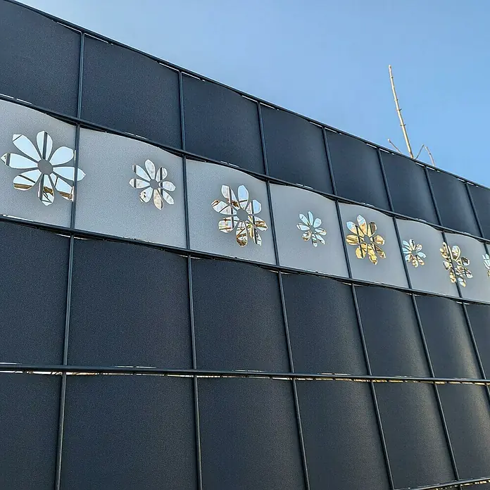 zaun|zu Sichtschutzstreifen Premium - 19 x 0,1 cm, Enzianblau, Sonnenblumen- Design - Detailansicht eines Zauns mit dekorativen, ausgestanzten Blumenmotiven in einer hellen, horizontalen Bahn. Der Zaun ist ansonsten dunkel und schlicht gehalten.