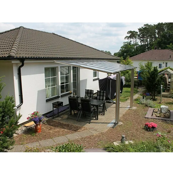Ximax Carport LINEA 80 - Außenmaß inkl. Dachüberstand (B x T): 2,73 x 4,95 m, Einzelcarport, Edelstahloptik - Ein Terrassendach aus Glas und Edelstahl bietet Schutz über einer Sitzgruppe im Garten. Das Haus mit braunem Dach grenzt an.