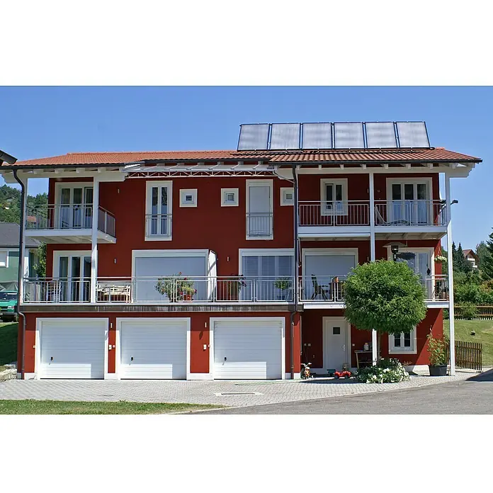 Sunset Solaranlage Sunsol 8 HZ/2 - Mit Frischwasserspeicher 825 l, Kollektorfläche: 15,06 m², Geeignet für: 3 - 6 Personen (Einfamilien- oder Mehrfamilienhaus) - Rotes Mehrfamilienhaus mit weißen Garagentoren, Balkonen und Solaranlage auf dem Dach. Das Haus hat mehrere Fenster und ist von Grün umgeben.
