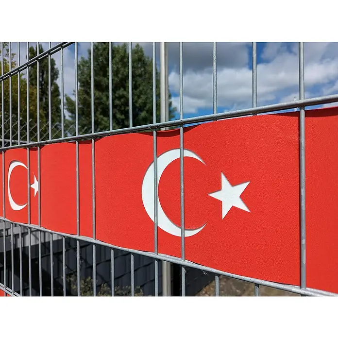 zaun|zu Sichtschutzstreifen Türkei - L x B x H: 2,55 m x 19 cm x 0,3 cm, Türkei, 3 Für 2 - Nahaufnahme einer Zauninstallation mit der türkischen Flagge, die durch das Gitter sichtbar ist und die Farben und Symbole des Landes zeigt.