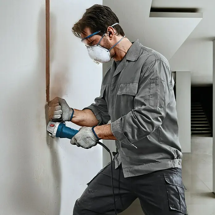 Bosch Professional Elektro-Winkelschleifer GWS 750 - 125 mm, Leerlaufdrehzahl: 11.000 U/min - Ein Bauarbeiter mit Schutzmaske und Brille benutzt einen blauen Bosch Winkelschleifer, um eine Nut in eine Wand zu fräsen.