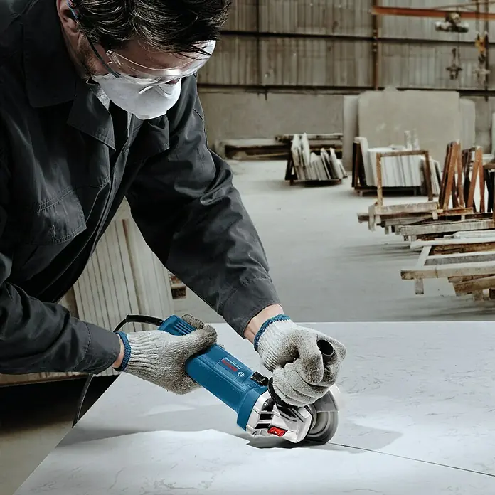 Bosch Professional Elektro-Winkelschleifer GWS 750 - 125 mm, Leerlaufdrehzahl: 11.000 U/min - Mann arbeitet mit einem blauen Bosch Winkelschleifer auf einer hellen Oberfläche, Schutzbrille und Maske inklusive.