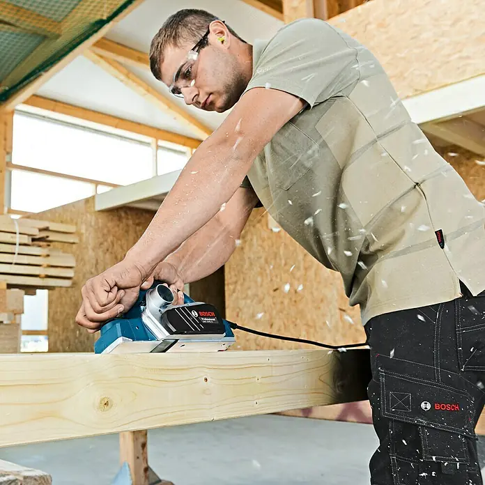 Bosch Professional Handhobel GHO 26-82 D - 710 W, Hobeltiefe: 0 mm - 2,6 mm, Leerlaufdrehzahl: 18.000 U/min - Ein Handwerker benutzt einen Bosch Hobel, um ein Holzbrett zu bearbeiten. Späne fliegen, während er das Werkzeug führt.